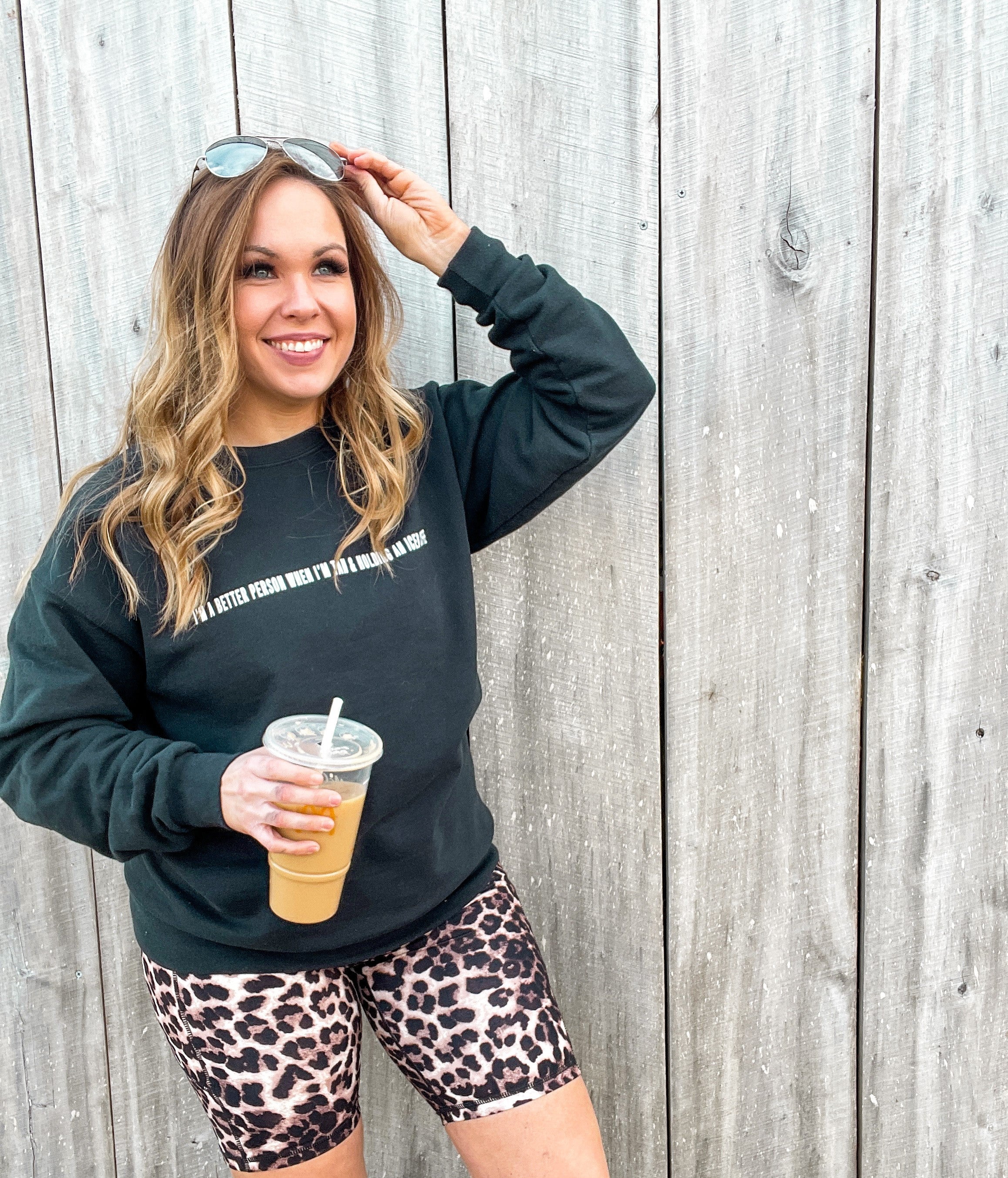 midsize influencer wearing black crewneck sweatshirt with I'm a better person when i'm tan and holding an iced coffee design in white letters